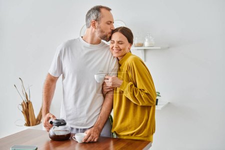 Mann küsst Kopf der fröhlichen Frau in der Nähe von frischem aromatischen Kaffee in Küche, kinderfreies Paar