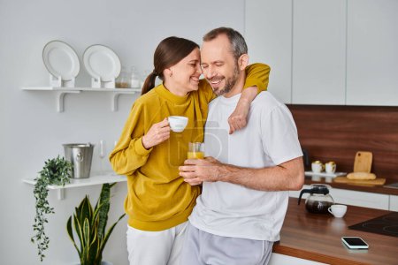 delighted child-free couple with coffee and fresh orange juice smiling with closed eyes in kitchen