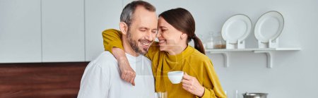 loving woman with cup of morning coffee embracing smiling husband in kitchen, horizontal banner