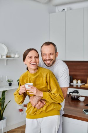 cheerful man embracing laughing wife holding fresh morning coffee in kitchen, child-free concept