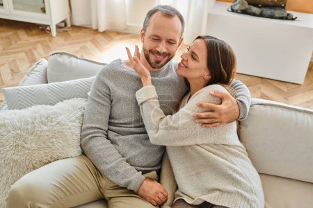 joyful woman embracing happy husband on cozy couch in living room, leisure of child-free couple