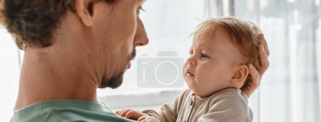 curly-haired and bearded father holding his infant baby boy in a cozy bedroom at home, banner