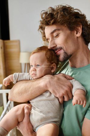curly-haired and cheerful father holding in arms his infant son in cozy bedroom at home, fatherhood