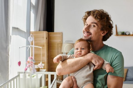 curly-haired and happy father holding in arms his baby son in cozy bedroom at home, fatherhood