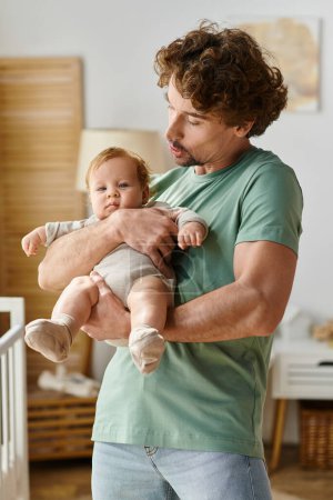hombre de pelo rizado y barbudo sosteniendo en brazos a su hijo bebé en el acogedor dormitorio en casa, la paternidad