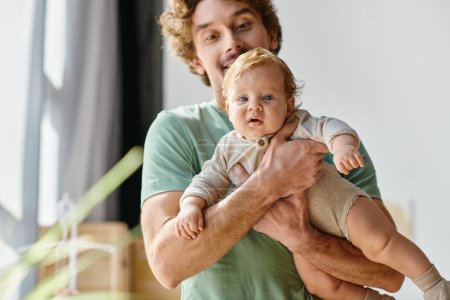 curly-haired and bearded man holding in arms his baby son with blue eyes at home, fatherhood