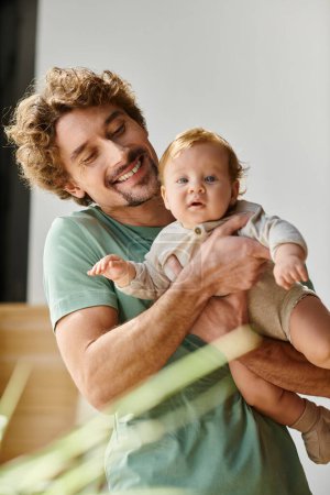 padre rizado y complacido sosteniendo en brazos a su hijo en el acogedor dormitorio en casa, la paternidad