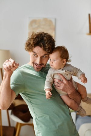 curly-haired father holding in arms his infant son and pointing away with finger in cozy bedroom