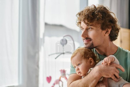 curly-haired and pleased father holding in arms his infant boy in cozy bedroom at home, fatherhood