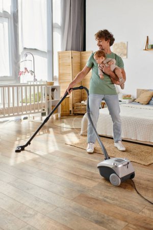 curly father multitasking housework and childcare, man vacuuming bedroom with infant son in arms
