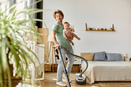 man multitasking housework and childcare, curly father vacuuming apartment with infant boy in arms