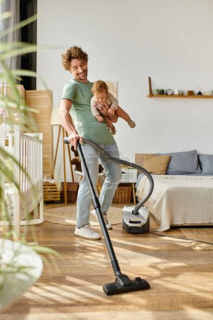 Mann Multitasking Hausarbeit und Kinderbetreuung, lächelnder Vater staubsaugt Wohnung mit Säugling im Arm