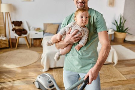 cropped man multitasking housework and childcare, happy father vacuuming house with son in arms