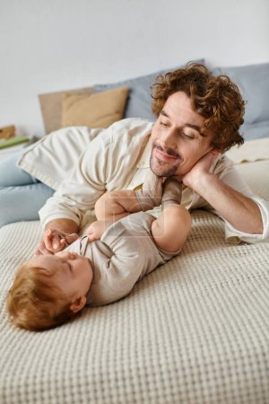 happy father with curly hair and beard looking at his baby son while lying together on bed, love