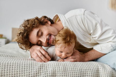 cheerful father with curly hair embracing his baby boy while lying together on bed, precious