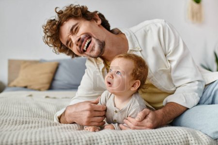 happy man with curly hair laughing while embracing his baby boy and lying together on bed