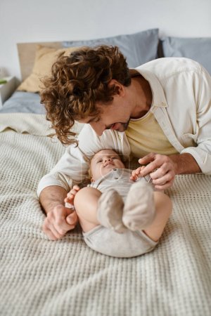 joyful father with curly hair and beard looking at his infant baby son on bed, precious moments