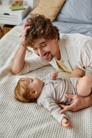 hombre alegre con el pelo rizado y la barba mirando a su bebé niño en la cama, momentos preciosos