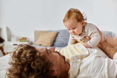 man with curly hair and beard playing with his infant son on a bed, bonding between father and child