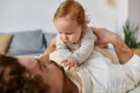 cute infant boy outstretching hands towards bearded dad on a bed, bonding between father and son