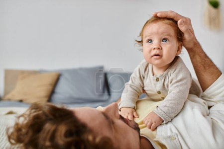 man with curly hair and beard stroking hair of with his infant son with blue eyes in bedroom