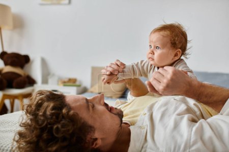 man with curly hair and beard holding hands of with his infant son with blue eyes in bedroom