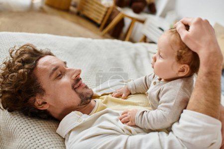 happy man with curly hair and beard stroking hair of with his infant son in bedroom, love and care