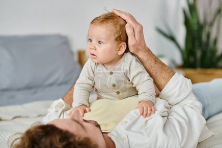 hombre con el pelo rizado abrazando a su hijo pequeño con ojos azules en una cama, vinculación entre el padre y el niño