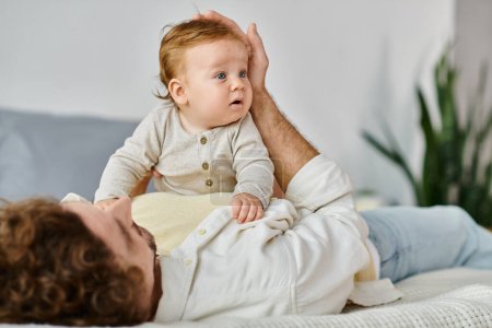 curly man lying on bed with his cute infant son with blue eyes, bonding between father and child