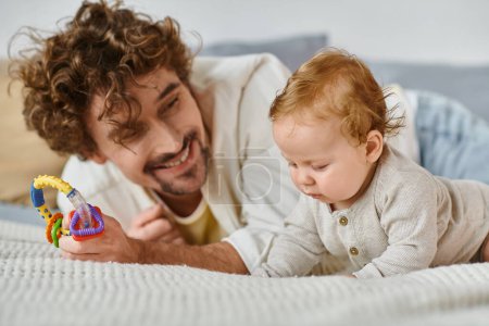 padre soltero sosteniendo sonajero colorido cerca bebé niño en el dormitorio, vínculo entre padre e hijo