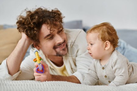 glücklicher Mann zeigt dem kleinen Jungen bunte Rassel im Schlafzimmer, Verbindung zwischen Vater und Sohn