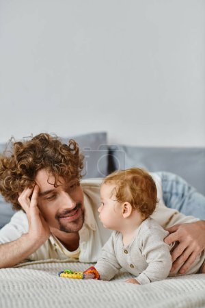 man looking at his infant baby boy near colorful rattle in bedroom, bond between father and son