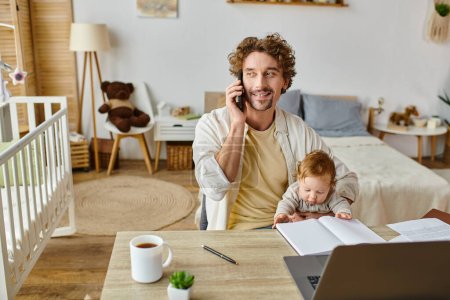happy man holding infant son while talking on smartphone and working from home, work-life balance