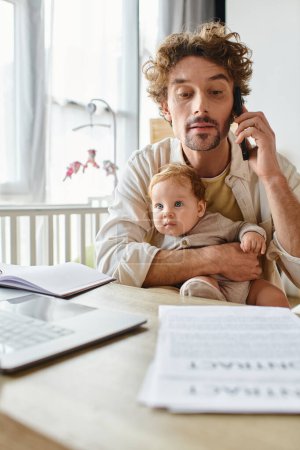 man holding infant son while talking on smartphone and working from home near papers and laptop