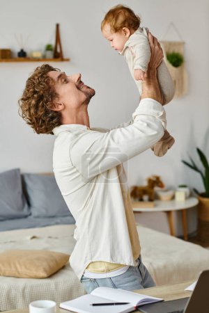 happy and curly father holding in arms his infant son near laptop and cup on desk, work life balance