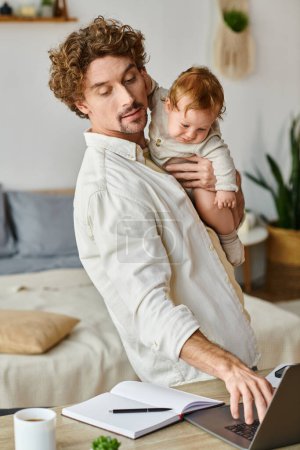 happy and curly father holding in arms his infant son and using laptop on desk, work life balance
