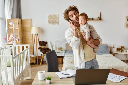 man holding infant son while talking on smartphone and looking at laptop on desk, work from home