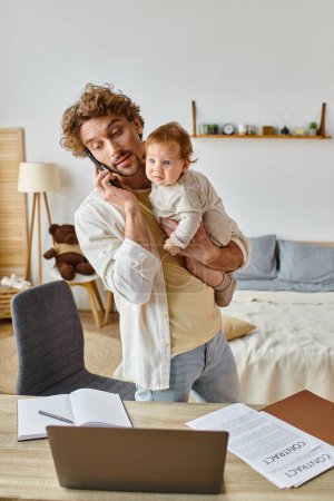 single father holding infant son while talking on smartphone near laptop on desk, remote work