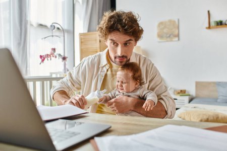 busy father holding baby bottle with milk and feeding infant son while working on project from home