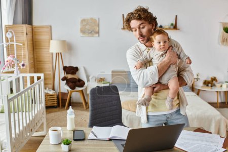 busy single father holding infant son and looking at papers near gadgets and baby bottle on desk