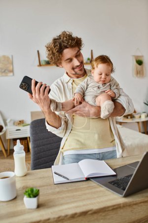 glücklicher alleinerziehender Vater hält Säugling Sohn und benutzt Smartphone in der Nähe von Laptop und Babyflasche auf Schreibtisch