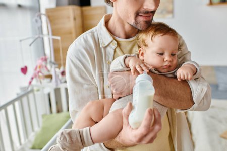 beschnitten Vater füttert seinen kleinen Sohn aufmerksam mit Milch in Babyflasche, Vaterschaft und Pflege