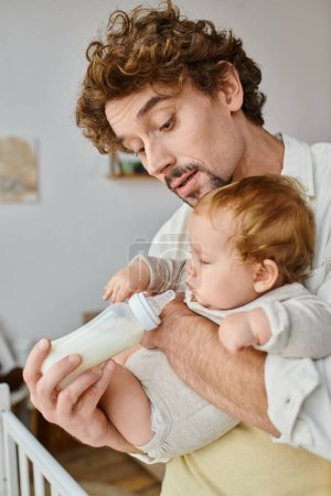 curly-haired father attentively feeding his infant son with milk in baby bottle, fatherhood and care