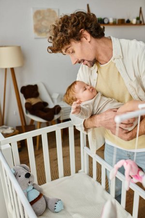 Photo for Curly single father holding in arms his infant son near crib in nursery, fatherhood and love - Royalty Free Image