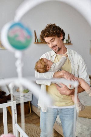 Photo for Happy single father holding in arms his infant son near crib in nursery, fatherhood and love - Royalty Free Image