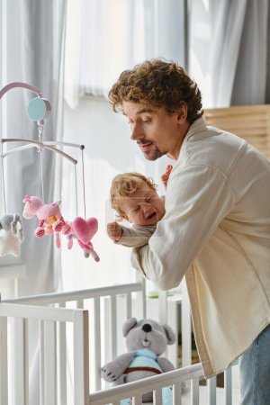 caring single father holding his crying infant son near baby crib in nursery, fatherhood and love