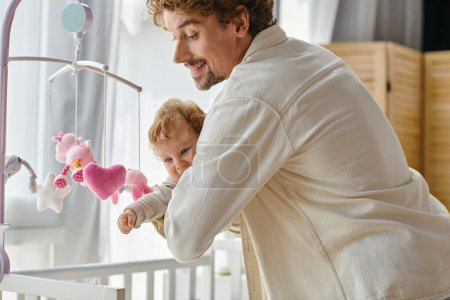 caring single father holding his infant son near baby mobile in nursery, fatherhood and love