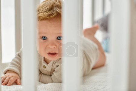 Niño bebé de ojos azules mirando a través de listones de la cuna en su cuarto de la guardería, la inocencia del niño