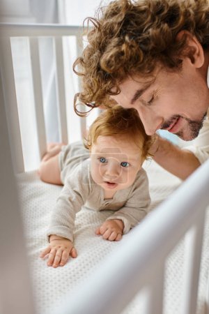 happy father with beard and curly hair embracing blue eyed baby boy in crib, parenthood and love