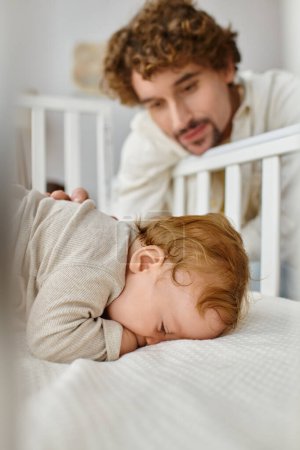niedliche Säugling Baby Junge schläft in der Krippe, während Vater ihn beobachtet, lediger Vater auf verschwommenem Hintergrund